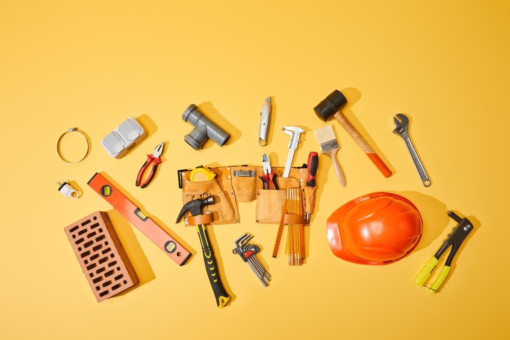 Ansicht von Werkzeuggürtel, Ziegel, Industriewerkzeug und Helm auf gelbem Hintergrund - Foto, Bild