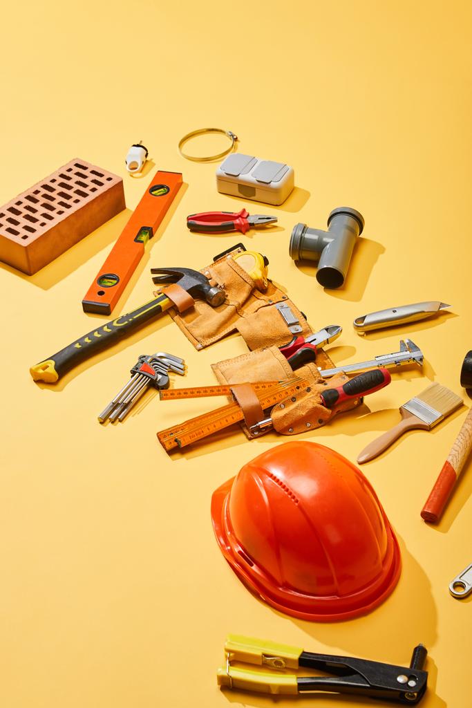hoge hoek weergave van gereedschapsriem, industrieel gereedschap, baksteen en helm op gele achtergrond - Foto, afbeelding