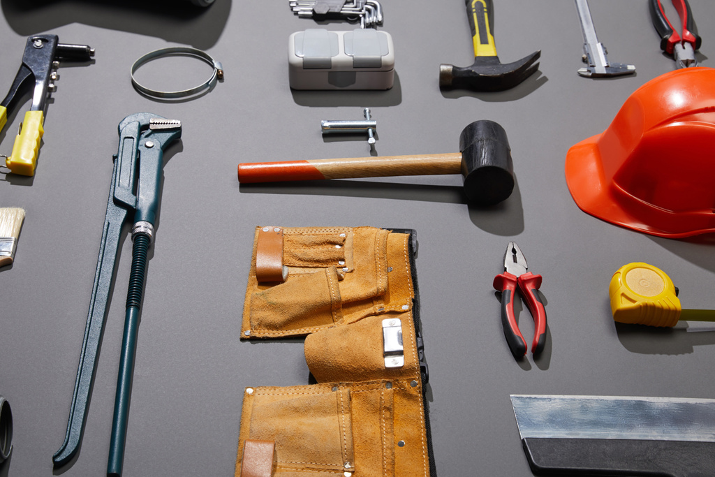 Blick auf Werkzeuggürtel, Helm, Hämmer, Affenschlüssel, Spachtelmesser, Zangen, Bremssättel, Nietpistole und Maßband auf grauem Hintergrund - Foto, Bild