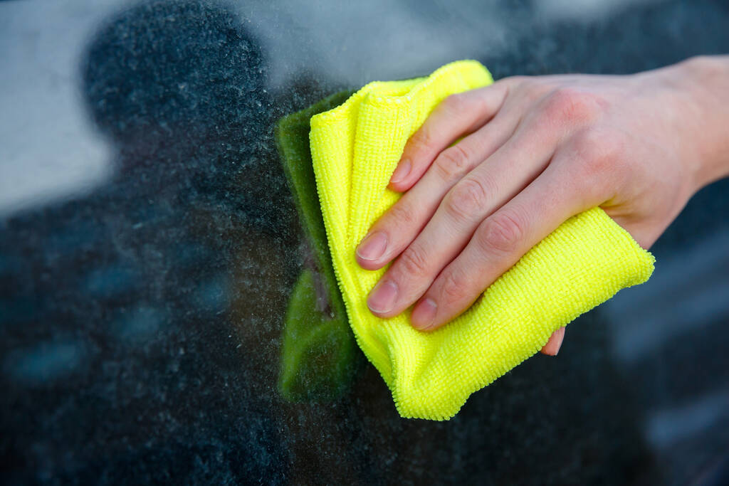 χέρι με ένα κίτρινο κουρέλι σκουπίζει το βρώμικο γυαλί ενός παραθύρου αυτοκινήτου - Φωτογραφία, εικόνα