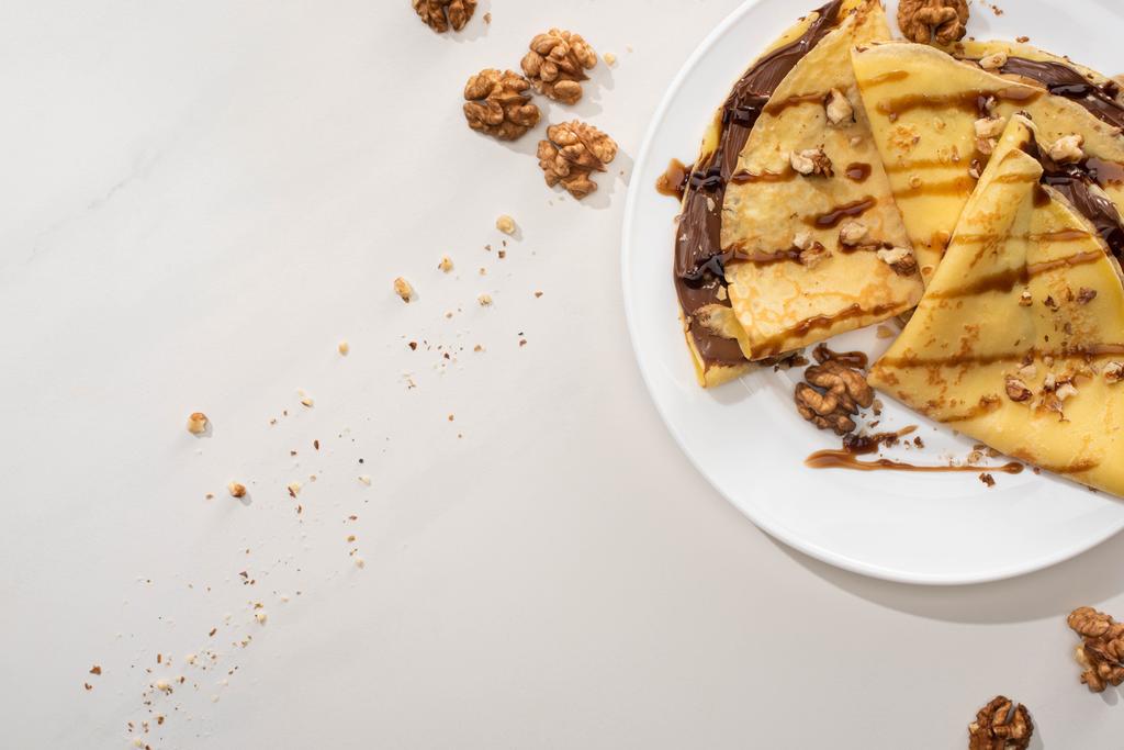 bovenaanzicht van lekkere crêpes met chocoladepasta en walnoten op plaat op grijze ondergrond - Foto, afbeelding