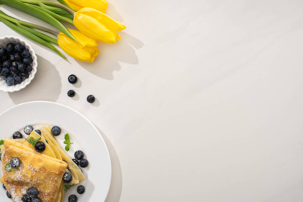 vrchní pohled na chutné palačinky s borůvkami a mátou na talíři v blízkosti žlutých tulipánů na šedém pozadí - Fotografie, Obrázek