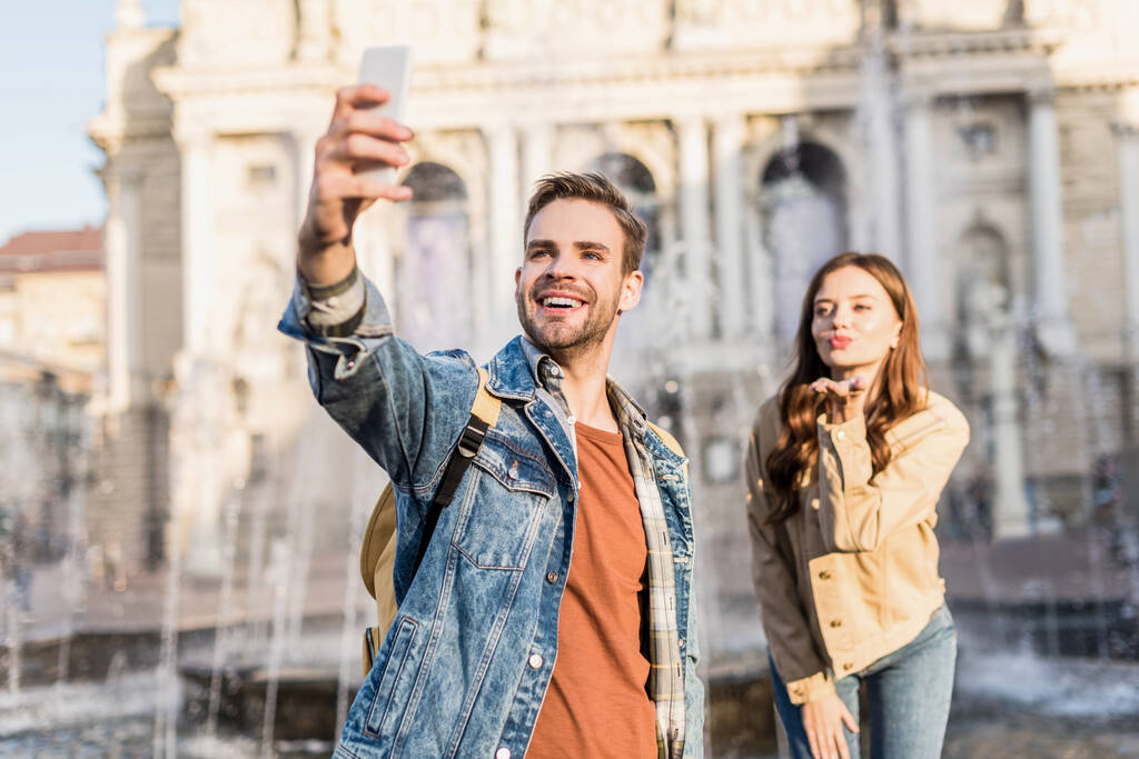 Selective focus of happy man with girlfriend blowing kiss taking selfie with smartphone near fountain in city - Photo, Image