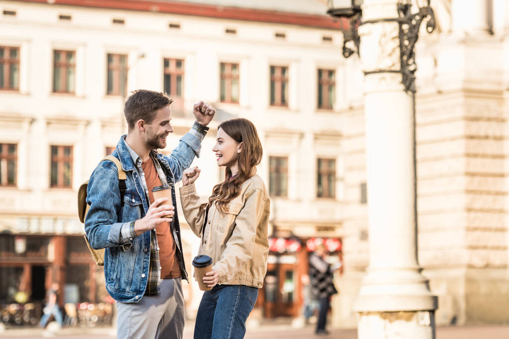 Emocionado novio y novia con tazas de café desechables mirándose y sonriendo en la ciudad
 - Foto, Imagen