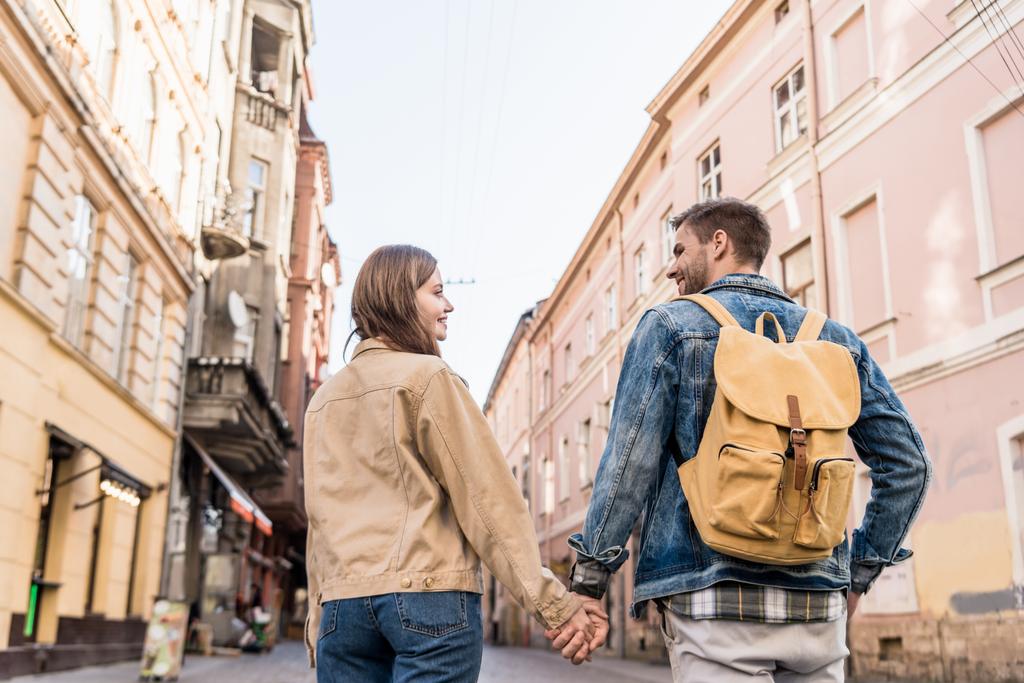 Back view of couple looking at each other and holding hands with backpack in city - Photo, Image