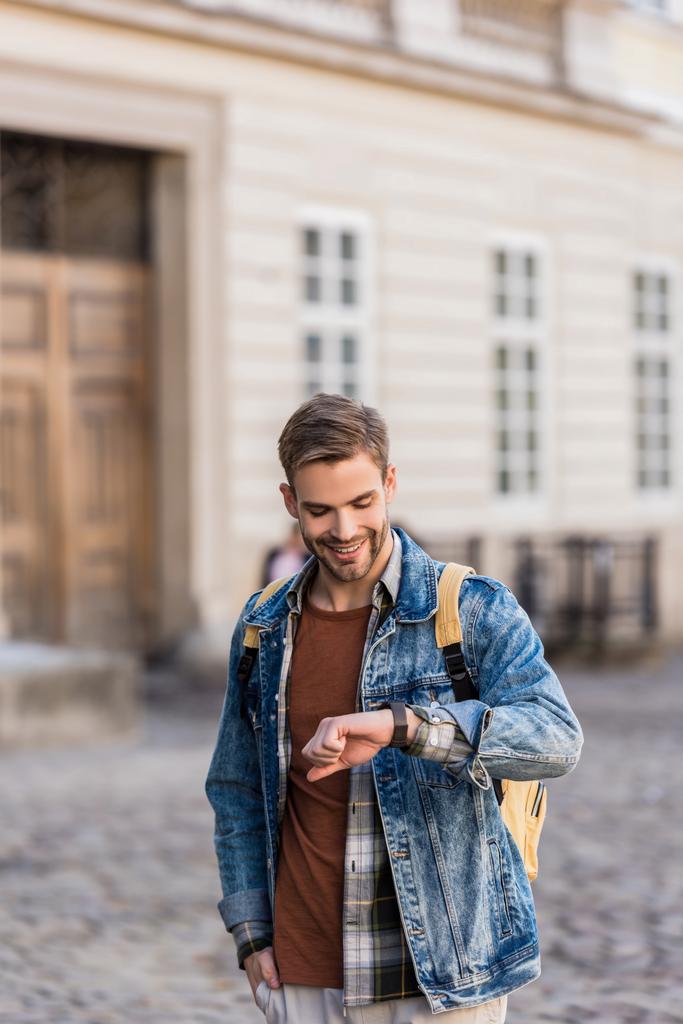Όμορφος άντρας με το χέρι στην τσέπη χαμογελώντας και κοιτάζοντας ρολόι χειρός στην πόλη  - Φωτογραφία, εικόνα