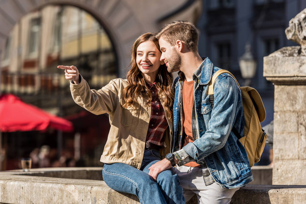 Petite amie pointant du doigt et regardant loin avec petit ami assis ensemble sur la pierre et souriant en ville
 - Photo, image