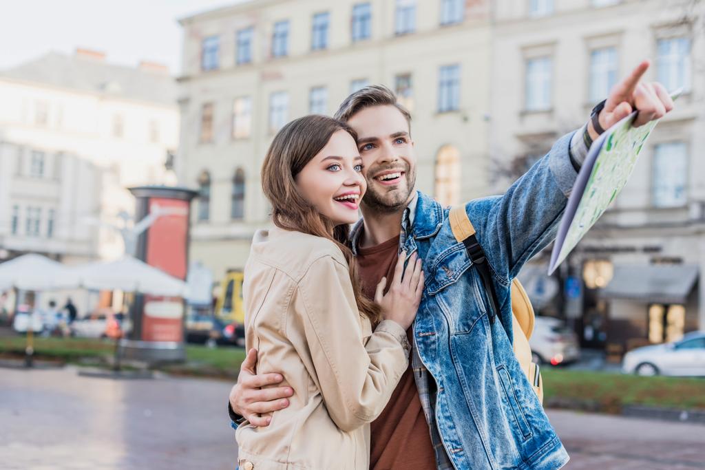 Muž se usmívá, drží mapu, ukazuje prstem a objímá šťastnou ženu ve městě - Fotografie, Obrázek