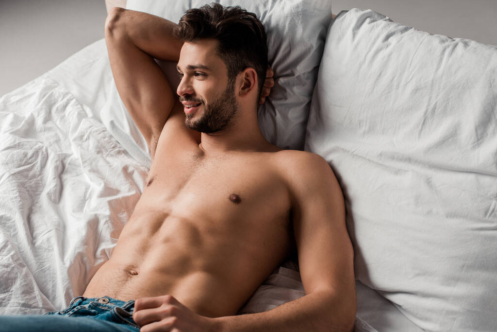 usměvavý sexy bez košile macho v džínách ležící na posteli na šedé - Fotografie, Obrázek