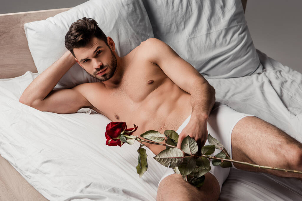 όμορφος άντρας ξαπλωμένος στο κρεβάτι με τριαντάφυλλο, απομονωμένος στο γκρι - Φωτογραφία, εικόνα