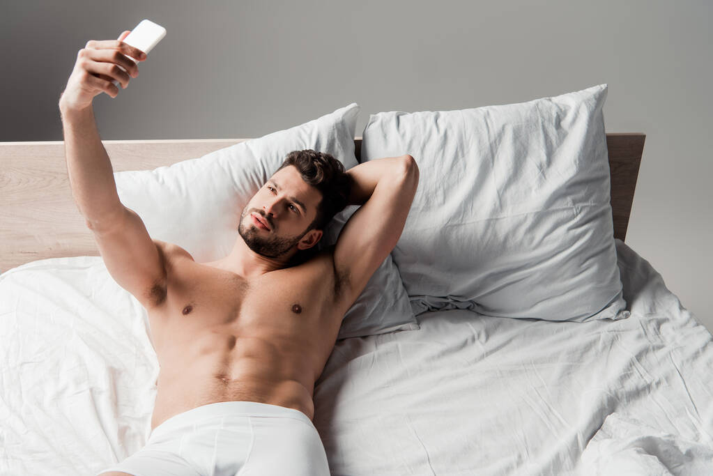 guapo sin camisa sexy hombre tomando selfie en teléfono inteligente en la cama en gris
 - Foto, Imagen
