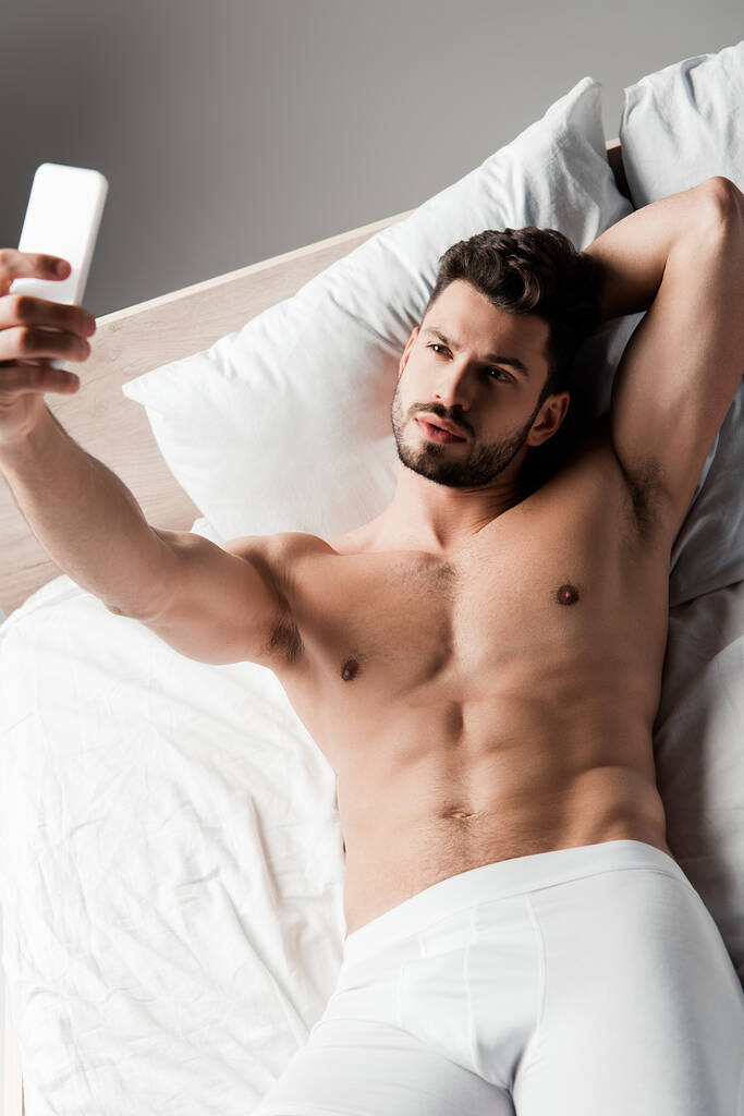 bez koszulki sexy macho biorąc selfie na smartfon w łóżku na szary - Zdjęcie, obraz