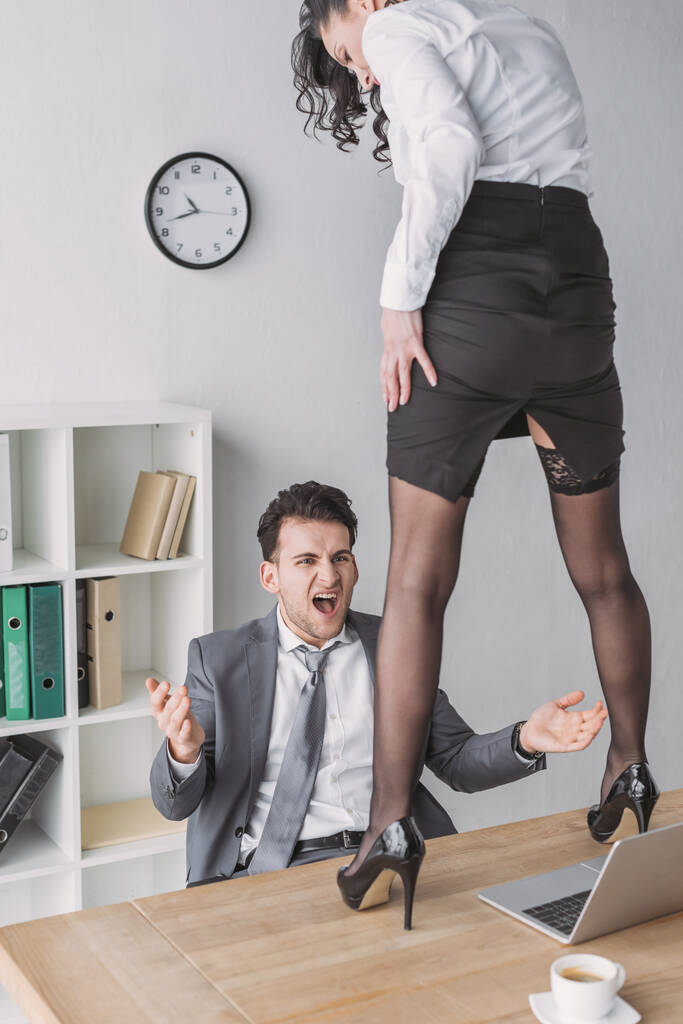 wütender Geschäftsmann schreit, während er die sexy Sekretärin ansieht, die in hochhackigen Schuhen auf dem Schreibtisch steht - Foto, Bild