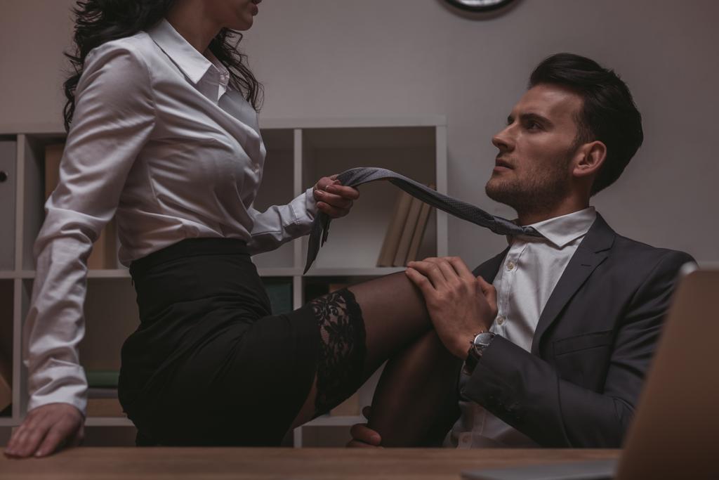 обрезанный вид соблазнительной секретарши, сидящей на столе и трогающей галстук красивого бизнесмена
 - Фото, изображение
