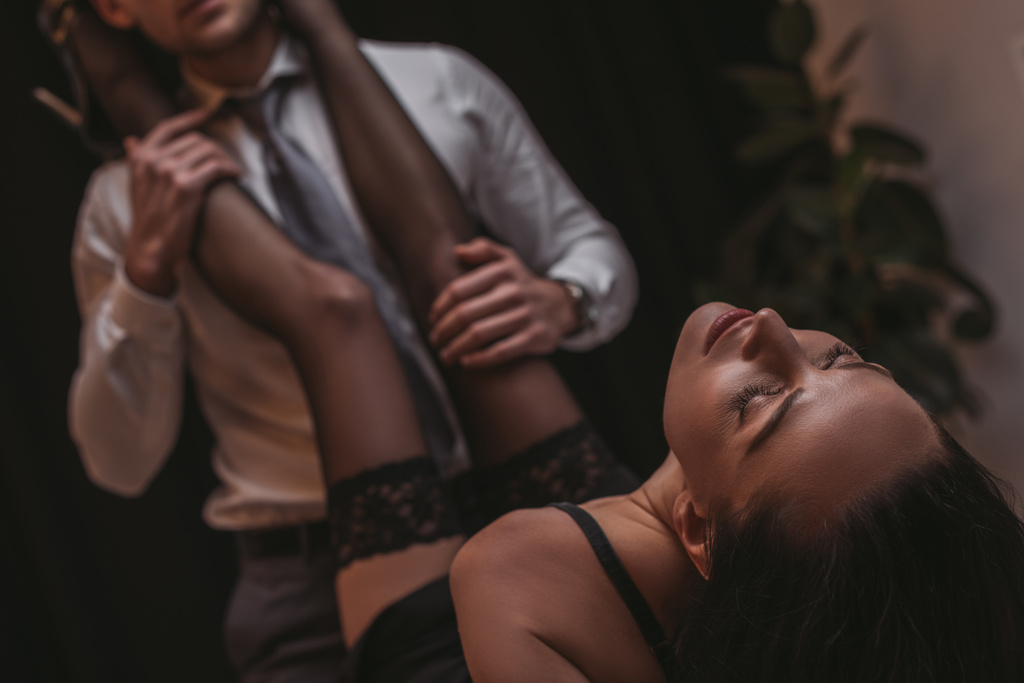 selective focus of businessman in formal wear touching legs of sexy secretary lying with closed eyes - Photo, Image