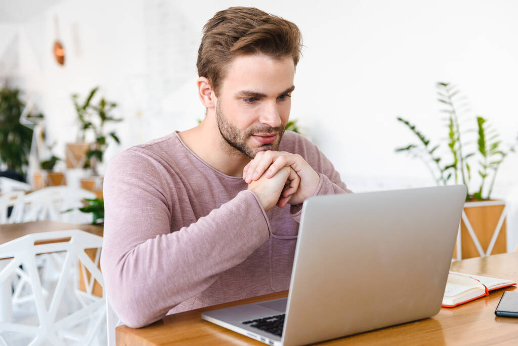 Obraz młodego brodatego mężczyzny pracującego na laptopie, siedzącego w kawiarni w pomieszczeniu - Zdjęcie, obraz
