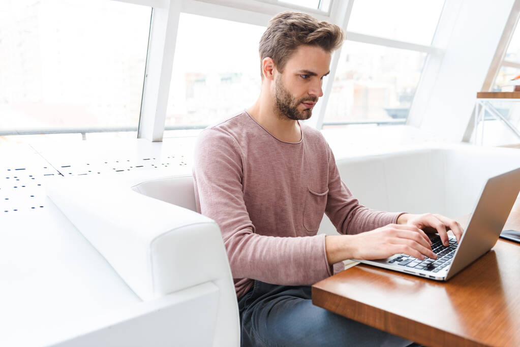 Imagen de un joven barbudo que trabaja en un ordenador portátil mientras está sentado en un café en el interior
 - Foto, imagen