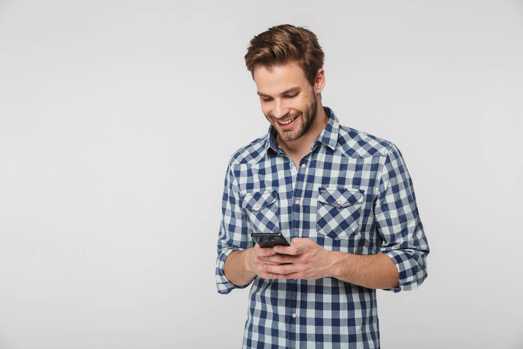 Ekose gömlek giyen, gülümseyen ve beyaz arka planda izole edilmiş cep telefonu kullanan mutlu genç adamın portresi. - Fotoğraf, Görsel