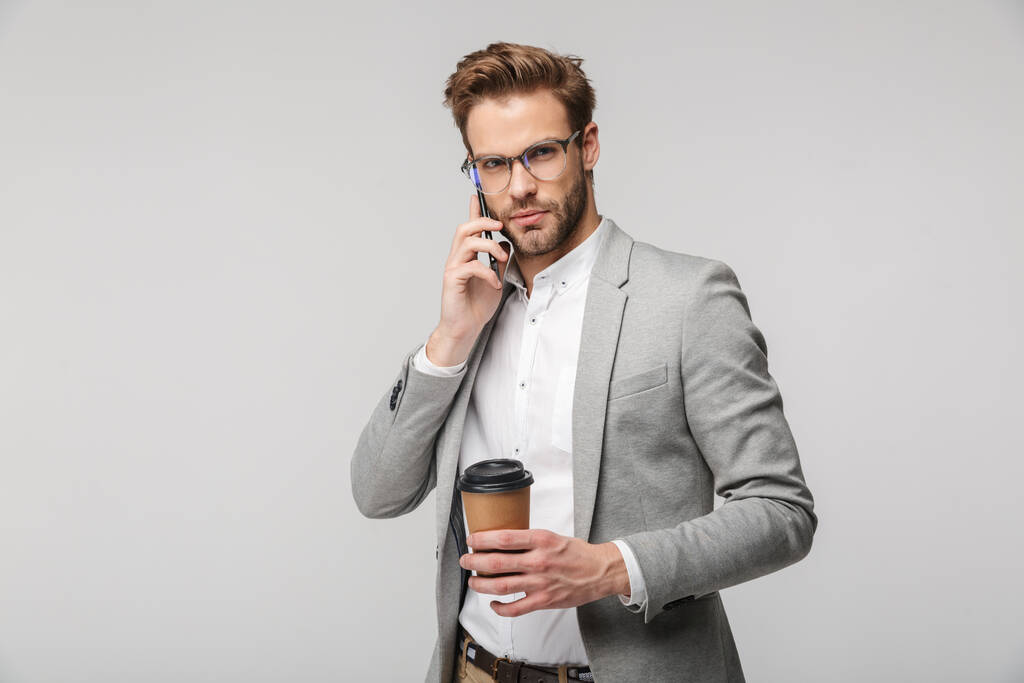Retrato de un hombre guapo serio con anteojos hablando por celular y sosteniendo una taza de papel aislada sobre fondo blanco
 - Foto, Imagen