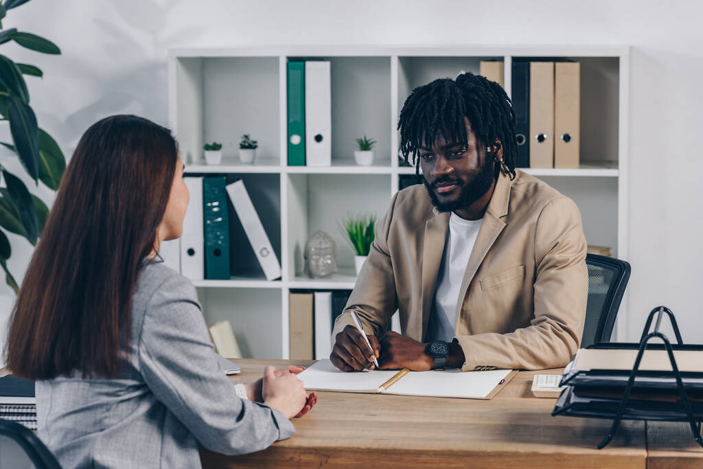 Reclutador afroamericano con bolígrafo y cuaderno realizando entrevista de trabajo con empleado en oficina
 - Foto, Imagen