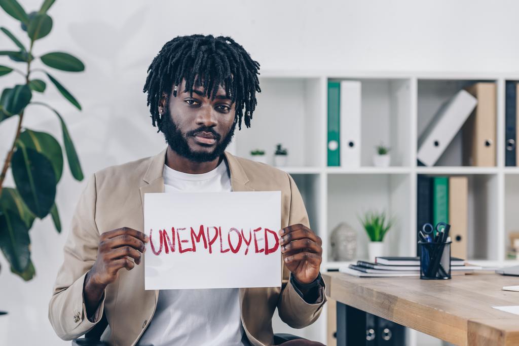 Impiegato afroamericano che guarda la macchina fotografica e presenta cartello con scritte disoccupate in ufficio
 - Foto, immagini