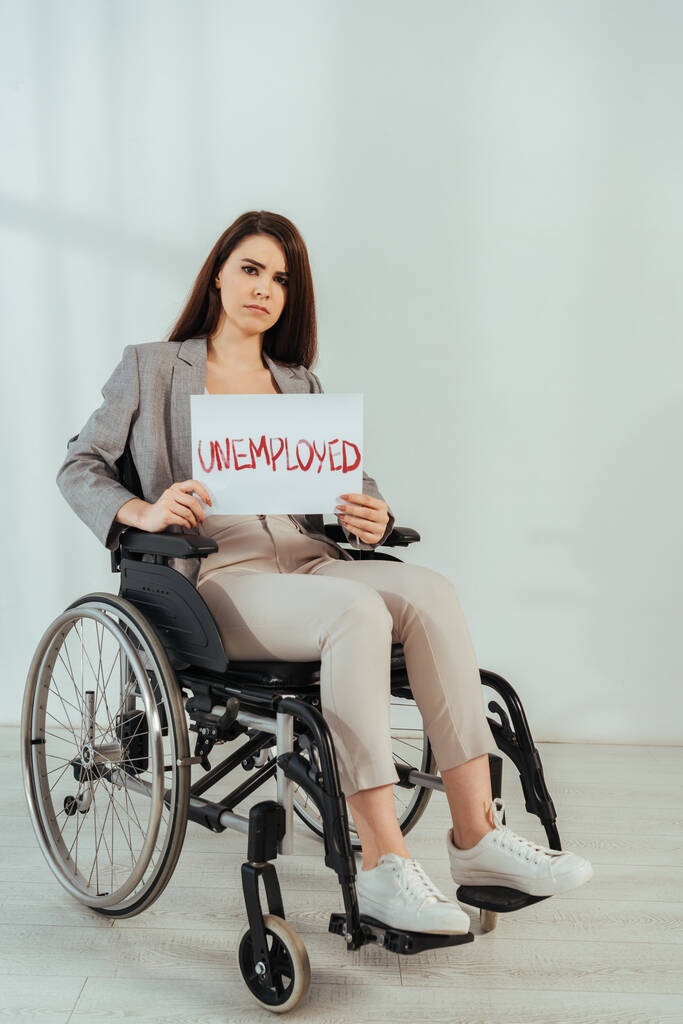 Mujer discapacitada mirando a la cámara y sosteniendo pancarta con letras desempleadas en silla de ruedas en blanco
  - Foto, Imagen