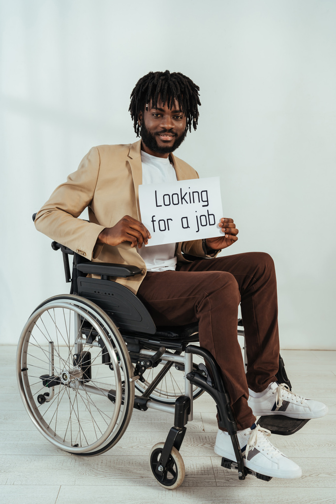 Zdravotně postižený africký Američan s plakátem s hledáním práce nápis na invalidním vozíku na bílém pozadí - Fotografie, Obrázek