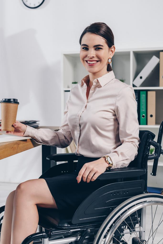 Employé handicapé en fauteuil roulant regardant la caméra et souriant avec une tasse de papier de café dans le bureau
 - Photo, image