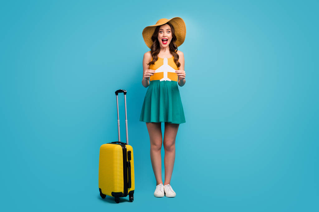 Full length photo of pretty lady traveler hold paper plane rolling suitcase luggage airport cheap flights wear sun specs short summer dress headwear shoes isolated blue background - Φωτογραφία, εικόνα