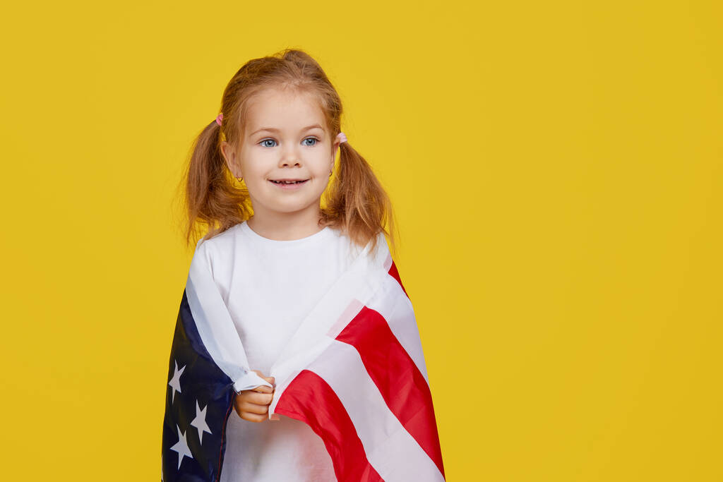 Hazafias ünnep. Boldog gyerek, aranyos kislány, amerikai zászlóval, sárga stúdió háttérrel. Az USA július 4-ét ünnepli, a függetlenség napját.. - Fotó, kép