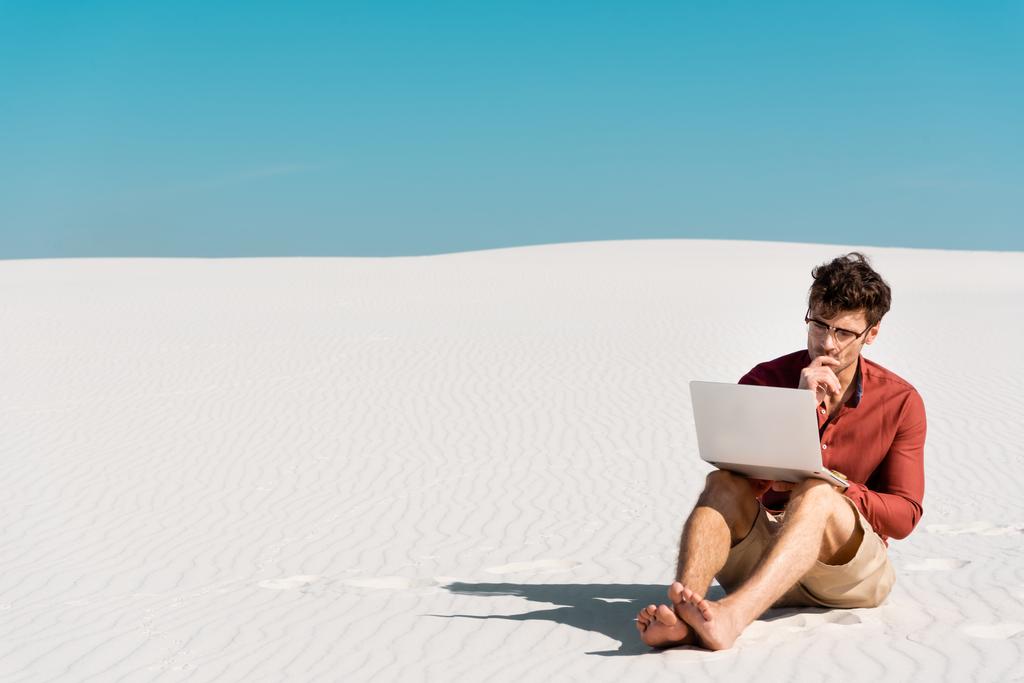 pensativo freelancer guapo en la playa de arena con portátil contra el cielo azul claro
 - Foto, Imagen