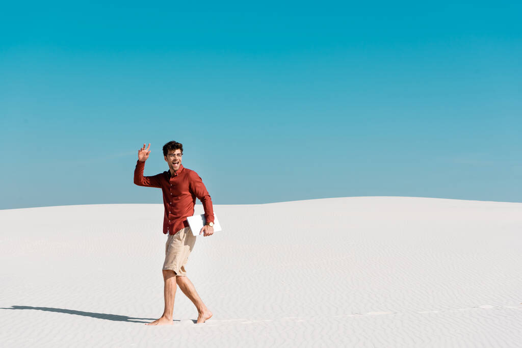 счастливый красавец-фрилансер на песчаном пляже с ноутбуком на фоне ясного голубого неба
 - Фото, изображение