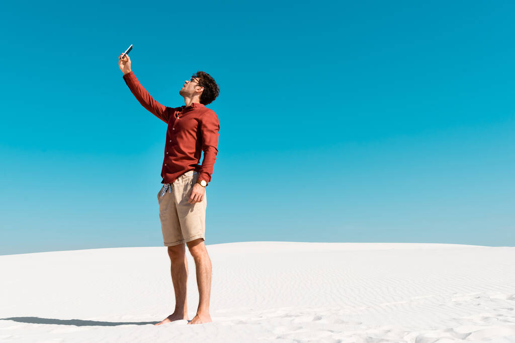 Kumsalda yakışıklı bir adam açık mavi gökyüzüne karşı akıllı telefondan konuşuyor. - Fotoğraf, Görsel