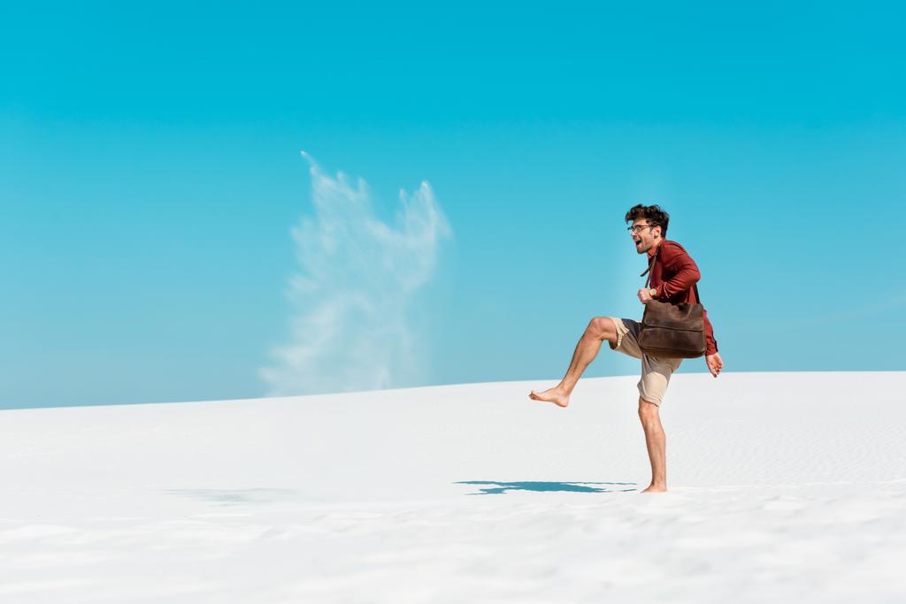 hombre feliz con bolsa de cuero lanzando arena en el aire en la playa
 - Foto, Imagen