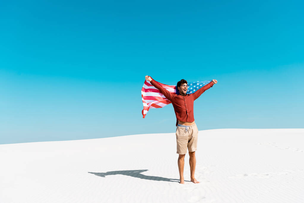 青い空を背景に風の強い砂浜にアメリカ国旗を掲げた幸せな男 - 写真・画像