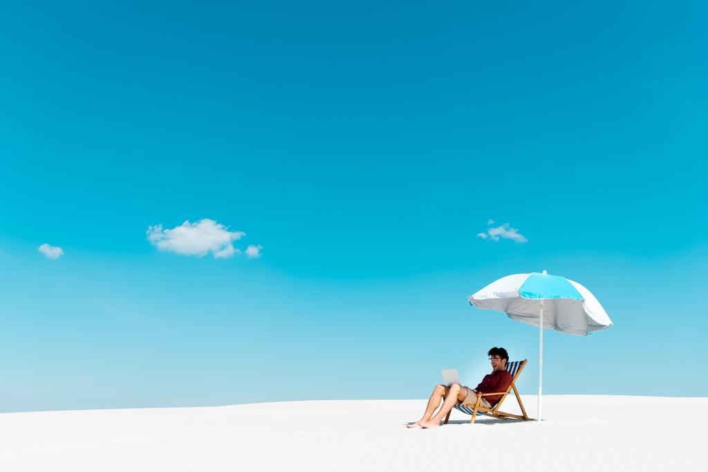 libero professionista seduto con computer portatile in sedia a sdraio sotto l'ombrellone sulla spiaggia sabbiosa contro il cielo blu
 - Foto, immagini