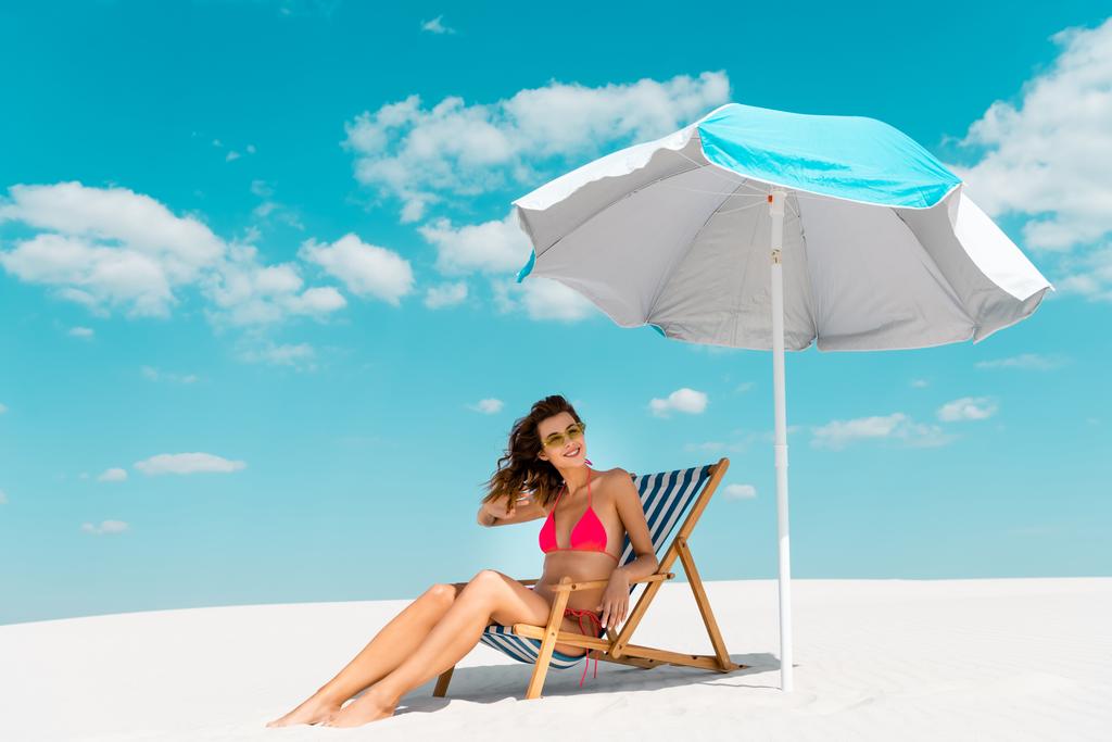 χαμογελαστή όμορφη σέξι κοπέλα με μαγιό και γυαλιά ηλίου κάθεται σε ξαπλώστρα κάτω από ομπρέλα στην αμμώδη παραλία - Φωτογραφία, εικόνα