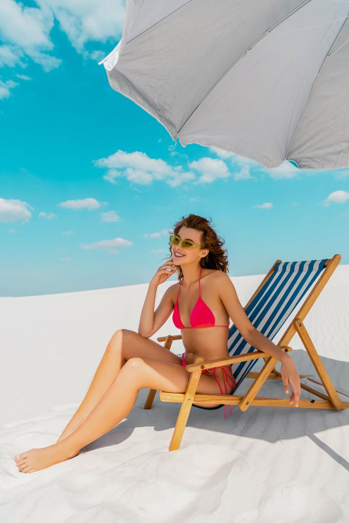 χαμογελαστή όμορφη σέξι κοπέλα με μαγιό και γυαλιά ηλίου κάθεται σε ξαπλώστρα κάτω από ομπρέλα στην αμμώδη παραλία - Φωτογραφία, εικόνα