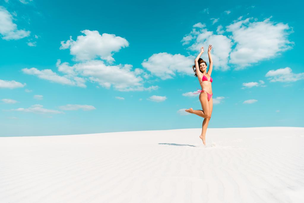 happy beautiful sexy girl in swimsuit jumping with hands in air on sandy beach with blue sky and clouds - Photo, Image