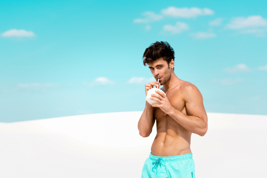 homem sexy com torso muscular em shorts de natação com bebida de coco na praia de areia
 - Foto, Imagem