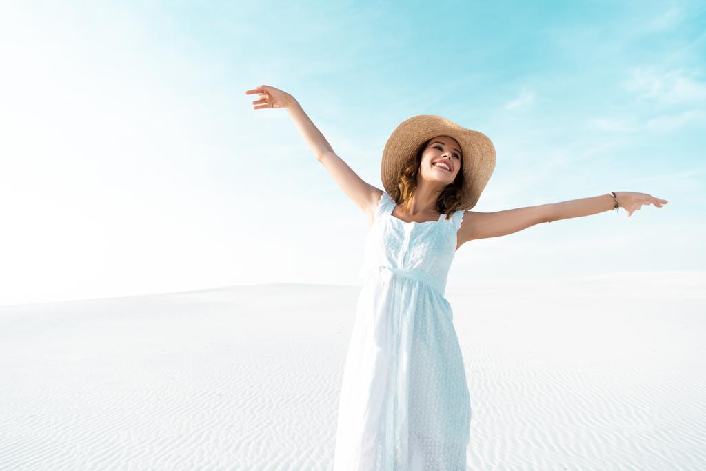 szczęśliwa piękna dziewczyna w białej sukience i słomkowym kapeluszu z wyciągniętymi rękami na piaszczystej plaży z błękitnym niebem - Zdjęcie, obraz