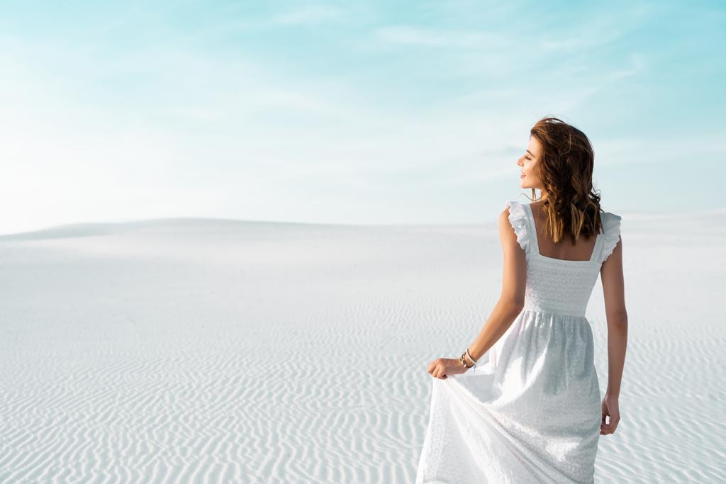 вид сзади красивой девушки в белом платье на песчаном пляже с голубым небом
 - Фото, изображение