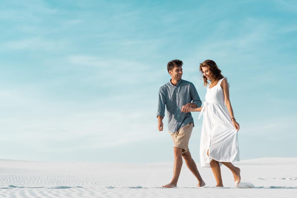 usmívající se mladý pár kráčející na písečné pláži proti modré obloze - Fotografie, Obrázek