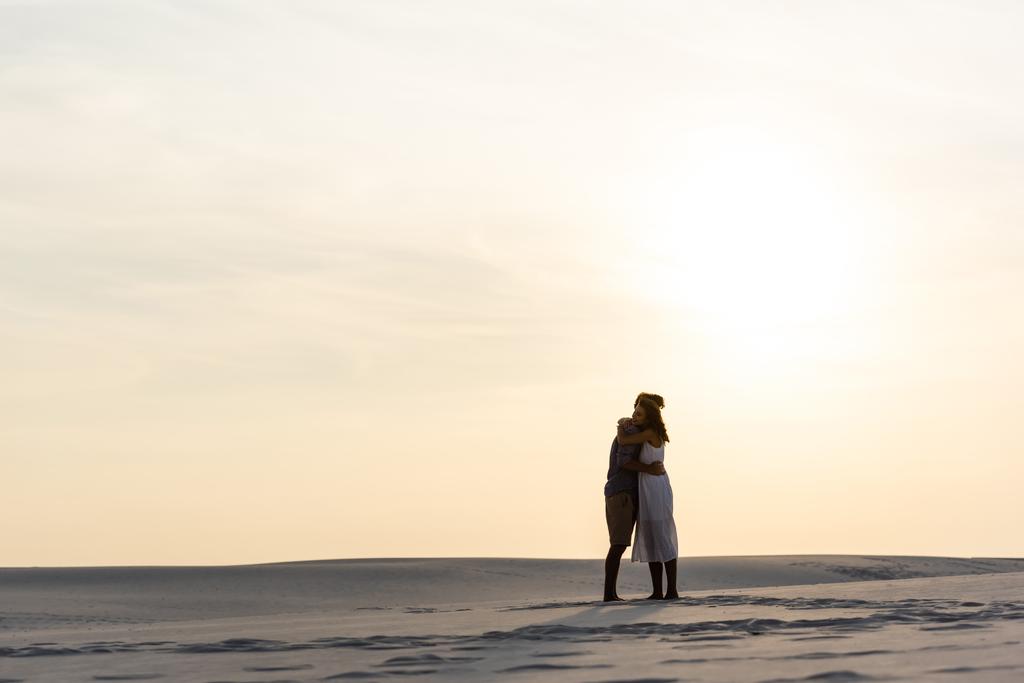 Seitenansicht eines jungen Paares, das sich bei Sonnenuntergang am Sandstrand umarmt - Foto, Bild