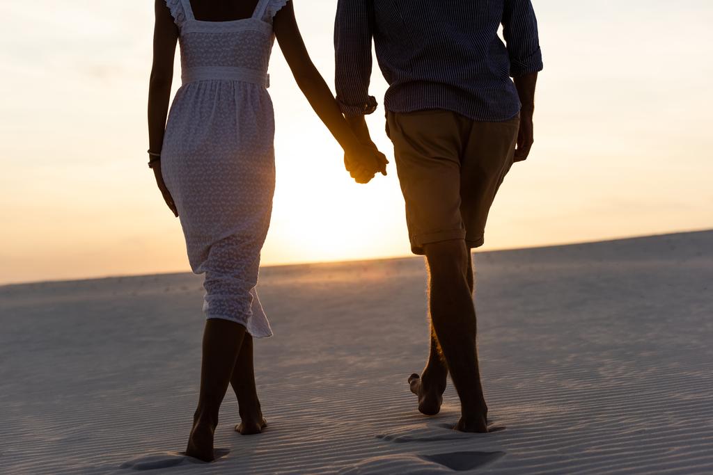vista cortada de homem e mulher de mãos dadas enquanto caminhava na praia contra o sol durante o pôr do sol
 - Foto, Imagem
