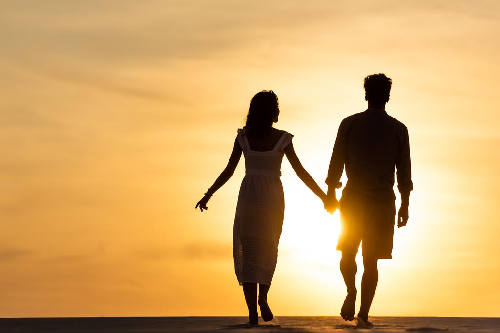 Silhouetten von Mann und Frau, die sich Händchen halten, während sie am Strand gegen die Sonne während des Sonnenuntergangs gehen - Foto, Bild