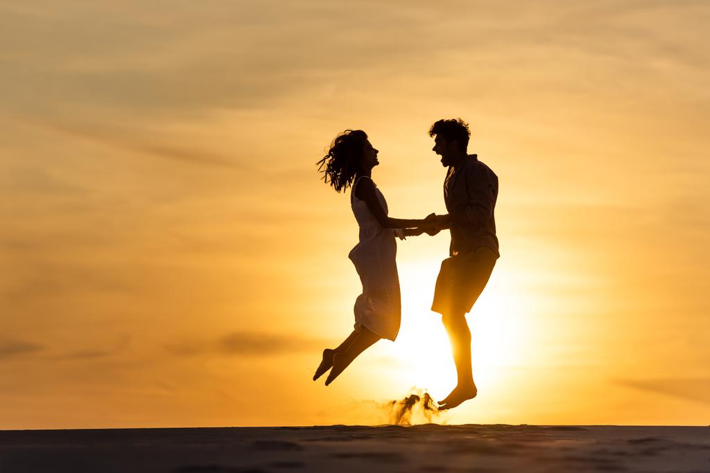 widok boczny sylwetki mężczyzny i kobiety skaczących na plaży przed słońcem podczas zachodu słońca - Zdjęcie, obraz