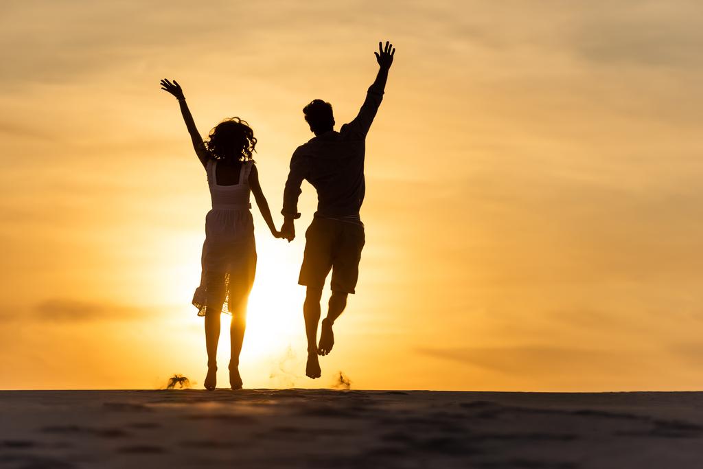 日没時に男と女のシルエットが太陽に向かってビーチに飛び込む - 写真・画像