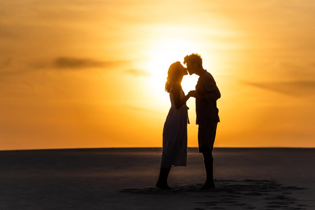 side view of silhouettes of man and woman kissing on beach against sun during sunset - Photo, Image