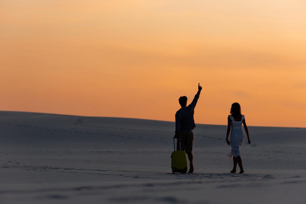 back view of couple walking on beach with raised hand and travel bag at sunset - Photo, Image